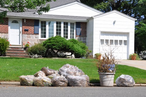 A residential property with a brick and white panel exterior, featuring a white garage door on the right and a bay window with multiple panes on the front facade. In the foreground, on the pavement near the curb, there is a collection of large, irregularly shaped rocks with a weathered appearance, some with visible moss or dirt. To the right of the rocks, a large grey plastic container holds a small, leafless shrub with thin, brown branches, and a few potted plants are placed nearby. The house is surrounded by a well-maintained green lawn with neatly trimmed bushes and a small tree. The scene appears to be captured during daytime in a suburban area, possibly in Wandsworth, with clear weather and bright natural light highlighting the textures of the stones and the smooth finish of the house’s exterior. This setting reflects an instance of rubbish or garden waste temporarily placed outside, potentially for collection or disposal by a waste removal service such as Commercial Waste Removal Wandsworth.