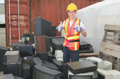 A male worker wearing a yellow safety helmet, a high-visibility orange and yellow vest, and purple gloves stands in an outdoor area surrounded by various discarded electronic appliances such as televisions, monitors, and computer equipment. The electronic waste, characterized by black, white, and dark grey plastic casings with glass screens, is stacked and spread across a paved surface, with some items leaning against a wooden pallet and others placed directly on the ground. In the background, there is a large reddish-brown shipping container and a white tarpaulin covering part of the area, indicating a waste removal or recycling site. The worker is smiling and giving two thumbs up, conveying a professional and positive attitude towards waste collection services. The scene is well-lit with natural daylight, highlighting the textures of the electronic waste and the uniformed worker, subtly reflecting the services offered by Commercial Waste Removal Wandsworth for handling bulky electronic rubbish in the Wandsworth area, inferred from the website context.