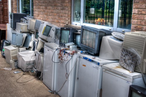 A row of discarded old computer monitors and desktop CPUs arranged outdoors on a concrete surface next to a brick building. The monitors include various types such as CRT (cathode-ray tube) and flat-screen displays, with some showing visible screens and others turned off. The materials include plastic casings, glass screens, and metal components, with the monitors exhibiting colors such as grey, beige, and black, and some showing signs of dirt or dust. Wires and cables are loosely draped over and among the equipment, with some disconnected or tangled. The background features a brick wall with a window that has a white frame and partially visible curtains, and green foliage reflected in the window. The setup appears to be part of an electronics waste disposal or clearance process possibly managed by Commercial Waste Removal Wandsworth, serving the Wandsworth area postcode in London, and situated in an outdoor environment adjacent to a commercial or residential building. The overall scene suggests a collection of obsolete IT equipment awaiting removal or recycling, consistent with rubbish disposal services in the local area.