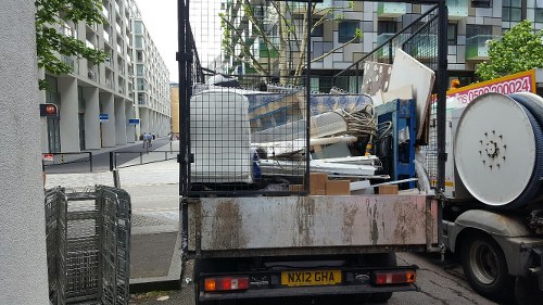 A small commercial waste collection truck is parked on a city street, with a metal cage loaded with various types of rubbish visible in the cargo area. The waste includes a mix of materials such as cardboard boxes, plastic containers, and metal objects, some stacked and leaning against the cage walls. The truck's body is weathered, showing signs of use with rust patches and dirt. The vehicle has a white wheel with a slightly worn tire visible on the right side, and the license plate reads 'NX12 GHA'. In the background, modern multi-storey residential buildings with balconies and large windows line the street, which has a crosswalk and traffic lights, indicating an urban setting possibly in Wandsworth. The scene is captured during daylight, with natural lighting highlighting both the vehicle and the surrounding environment, illustrating a typical rubbish removal operation conducted by Commercial Waste Removal Wandsworth in the area.