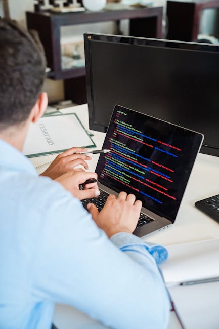 A person seated at a desk working on a laptop displays a screen filled with lines of colorful programming code, with a keyboard in front and a large computer monitor behind them. The workspace include