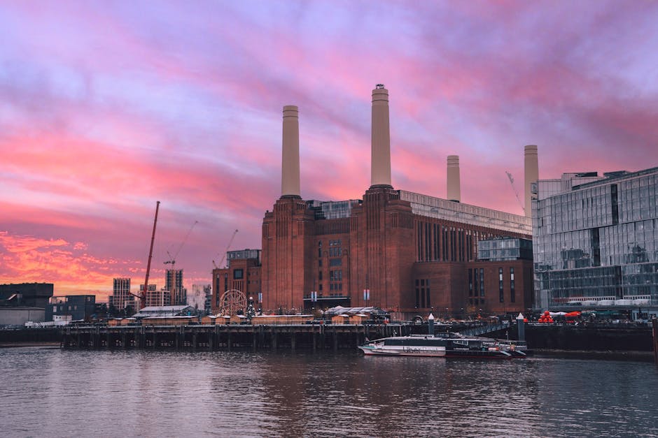 A view of the River Thames with a large, brown brick building featuring four tall chimneys in the background, alongside modern glass-fronted office buildings. In the foreground, part of a bridge or el